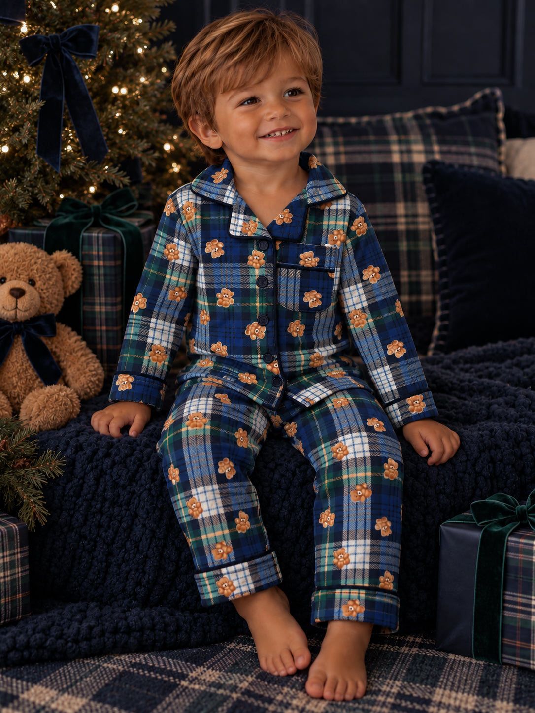 Smiling boy in bear-patterned pajamas, sitting on a cozy blanket near a Christmas tree and plush teddy bear.