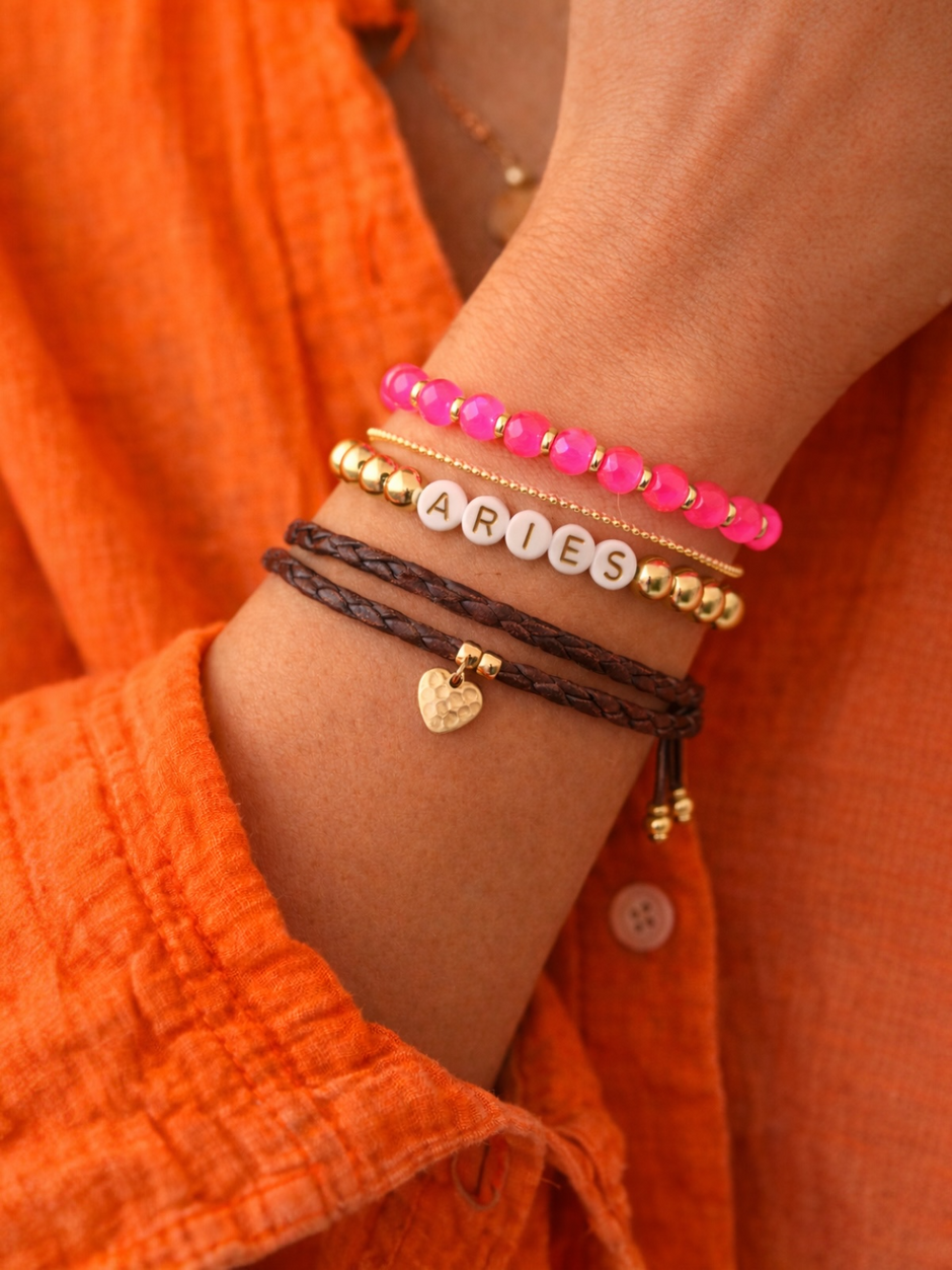 Close-up of a hand wearing stacked bracelets, including pink beads, gold accents, and an "Aries" charm.