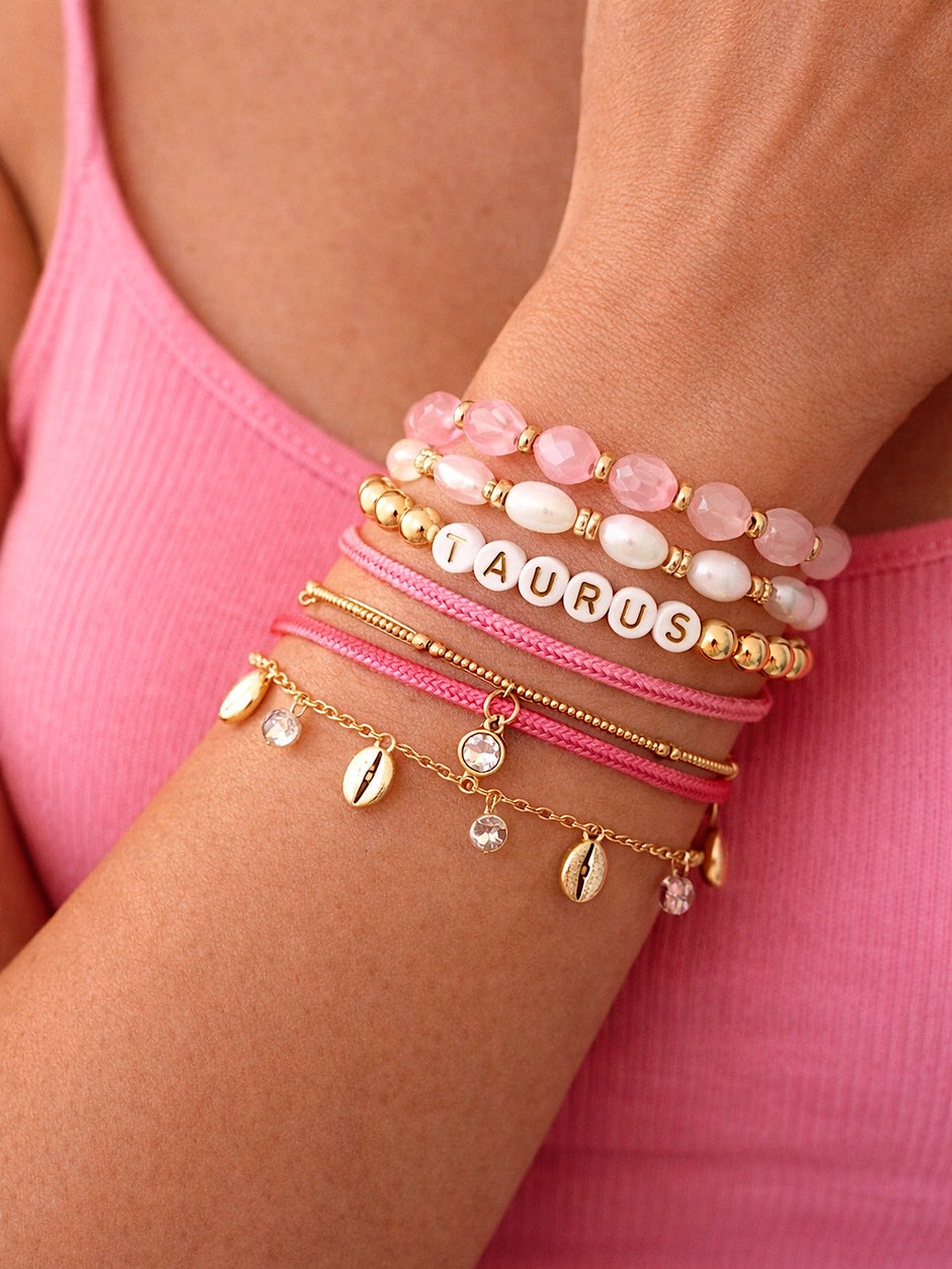 Close-up of a wrist adorned with various pink and gold beaded bracelets, featuring the word "Taurus."