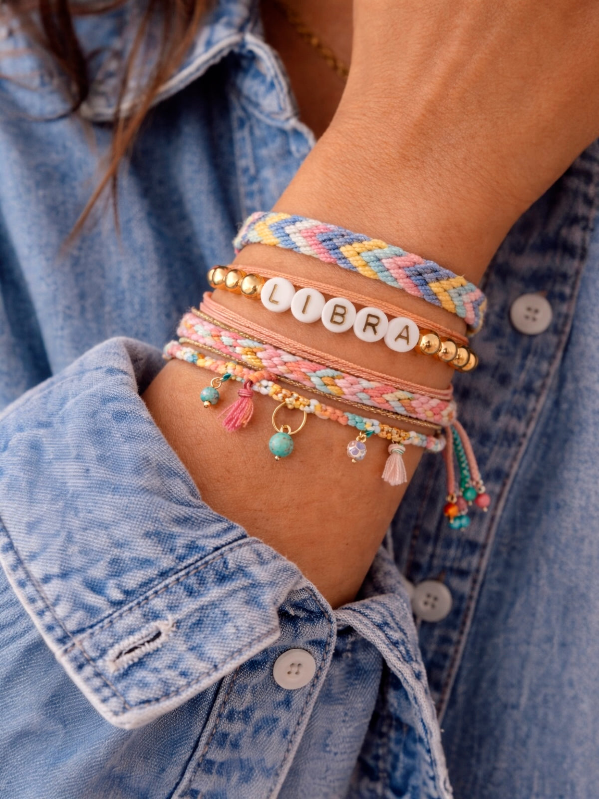 Colorful bracelets on a wrist, featuring beads, charm details, and "LIBRA" letters, against a blue denim shirt.