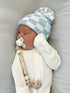 Newborn baby sleeping in a blue and white checkered hat, with a pacifier and a wooden name teether.