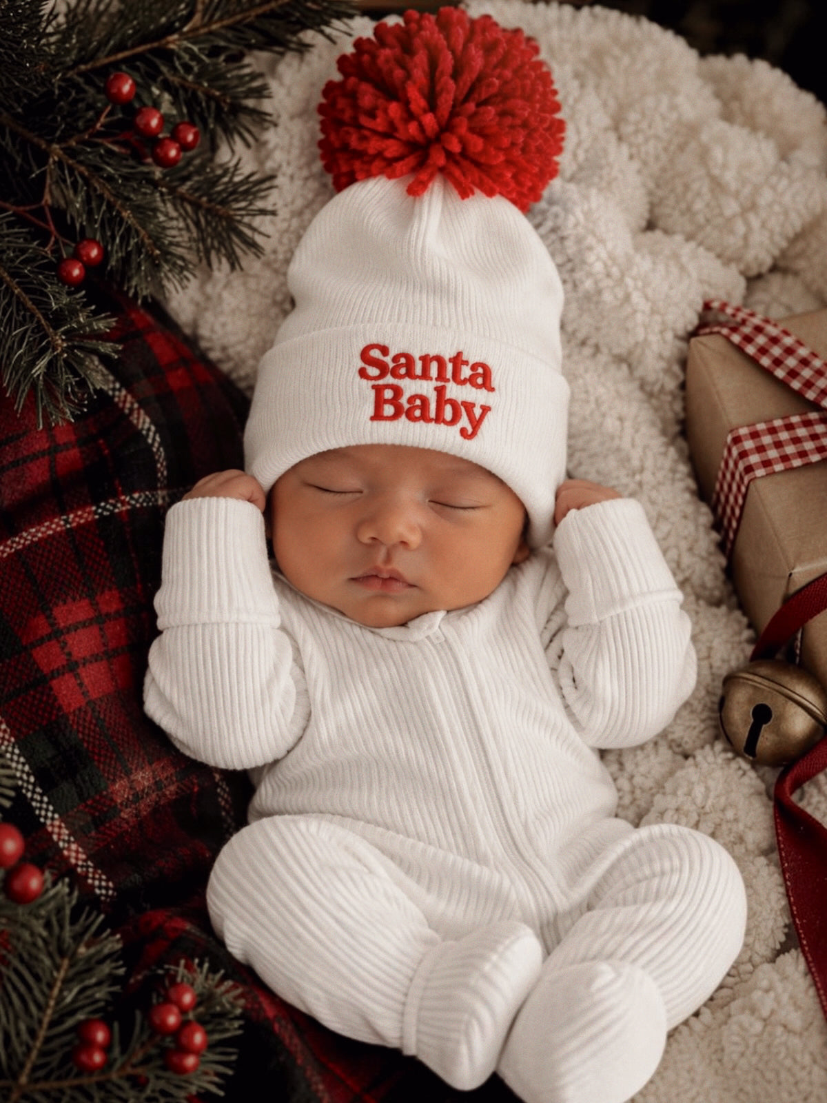 Baby sleeping in a cozy white outfit with a red pom-pom hat that says "Santa Baby," surrounded by festive decor.