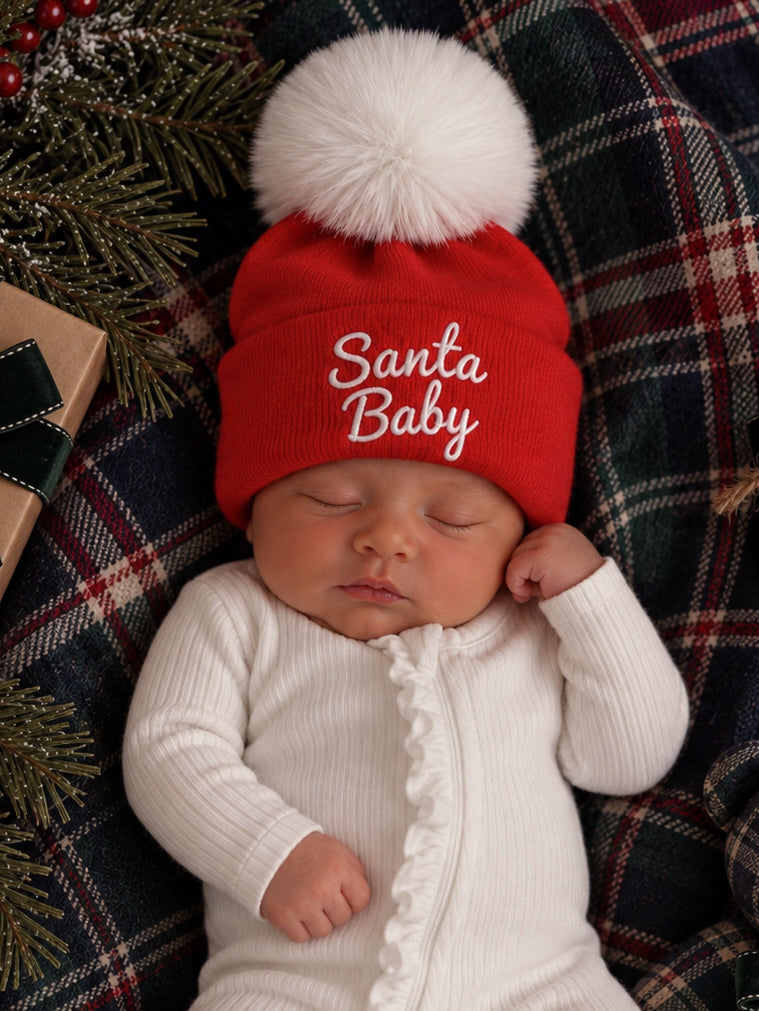 Infant wearing a red "Santa Baby" hat, lying on a plaid blanket with holiday decorations nearby.