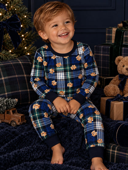 Smiling child in colorful plaid pajamas, sitting on a cozy blanket surrounded by holiday decor and gifts.