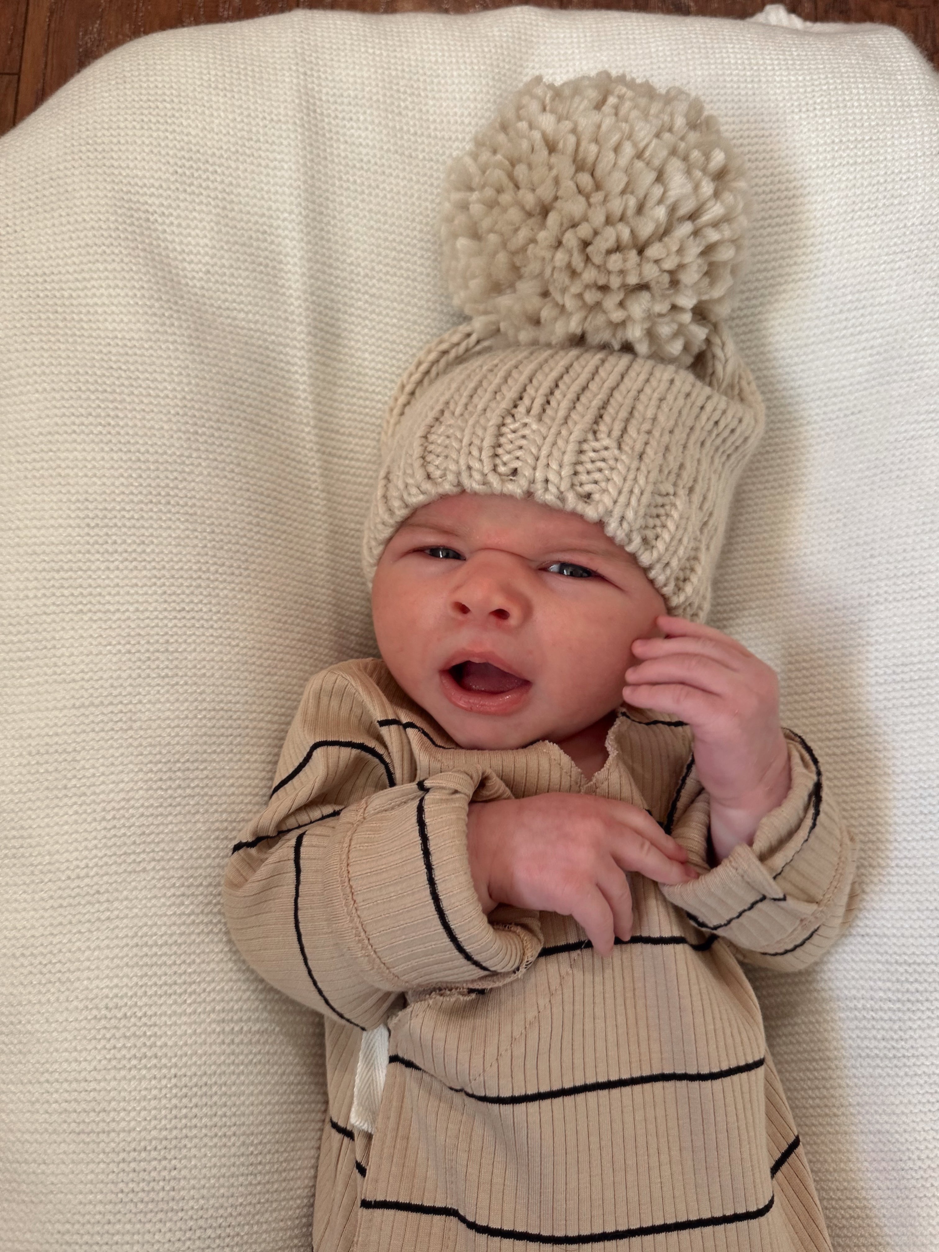 Newborn baby in a cozy beige hat with a pom-pom, wearing a striped outfit against a white background.