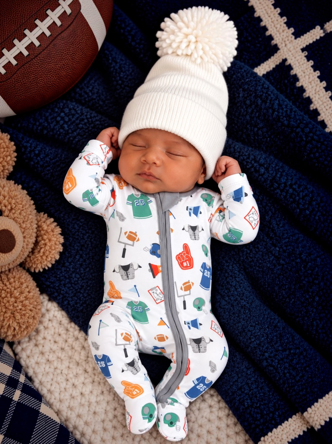 Baby sleeping on a cozy blanket, wearing a sports-themed onesie and a white pom-pom hat, with a teddy bear and football nearby.