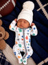 Baby sleeping on a cozy blanket, wearing a sports-themed onesie and a white pom-pom hat, with a teddy bear and football nearby.
