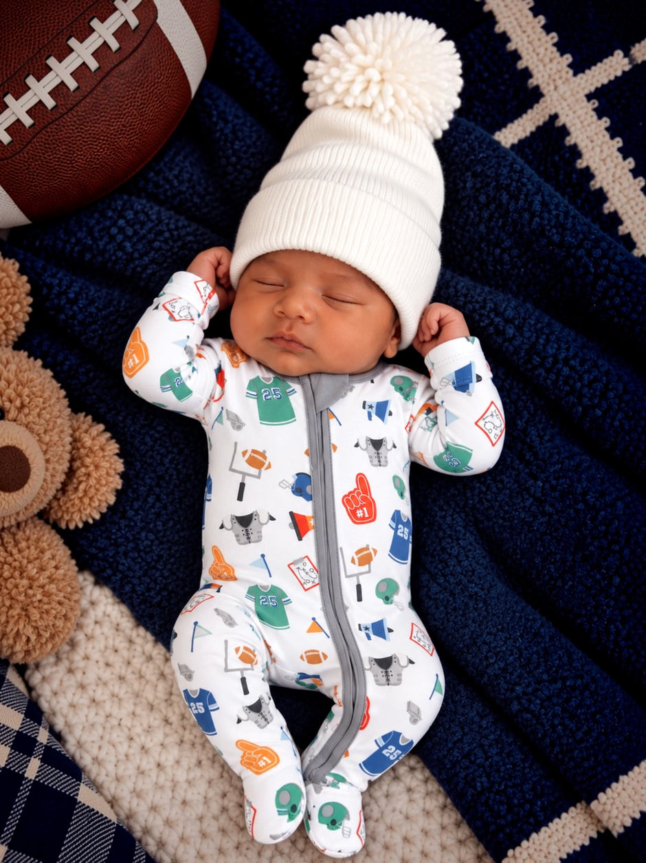 Baby sleeping on a cozy blanket, wearing a sports-themed onesie and a white pom-pom hat, with a teddy bear and football nearby.