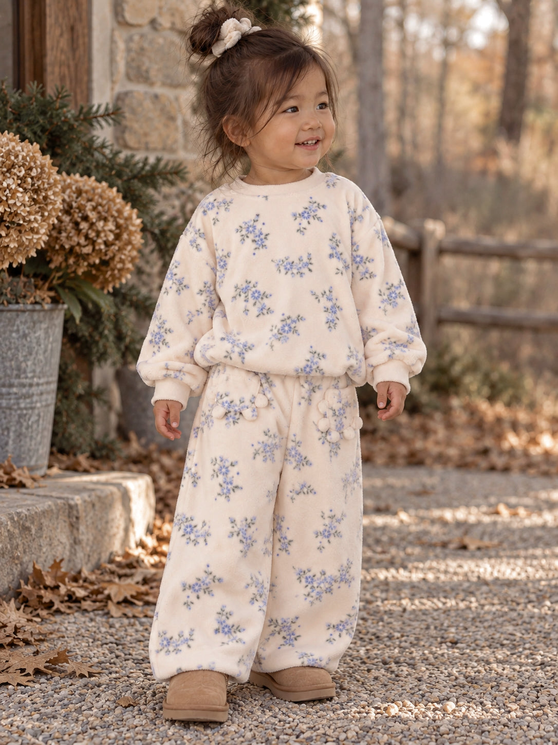 Child in floral loungewear smiles outdoors, surrounded by plants and autumn leaves. Cozy and comfortable outfit.