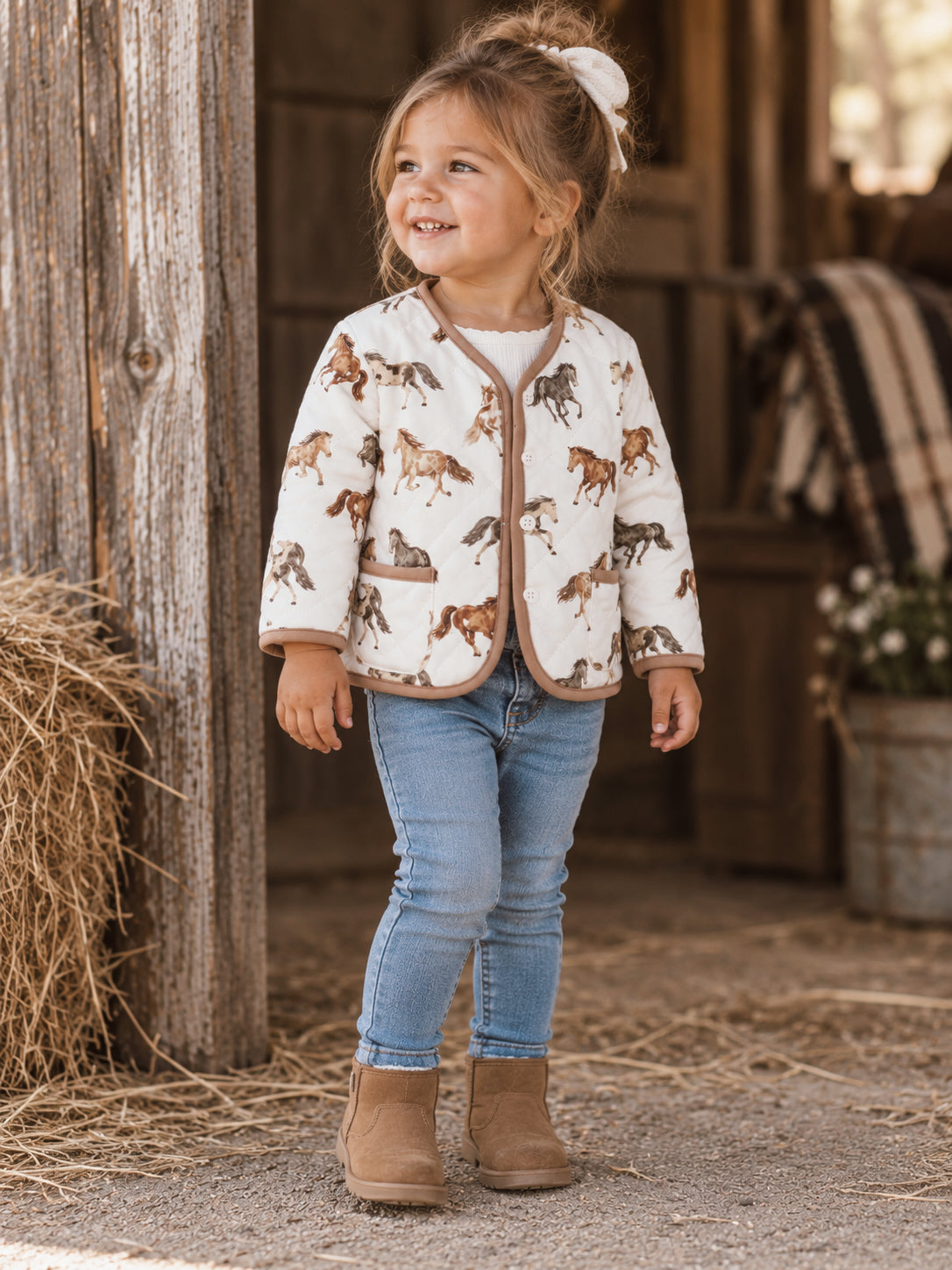 Smiling young girl in a horse-print jacket, jeans, and boots stands outdoors near hay bales, showcasing a playful expression.