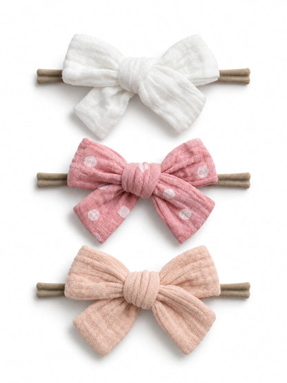 Three hair bows: white, pink with polka dots, and soft pink, all attached to brown hair elastics.