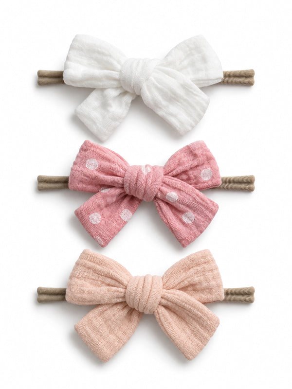 Three hair bows: white, pink with polka dots, and soft pink, all attached to brown hair elastics.