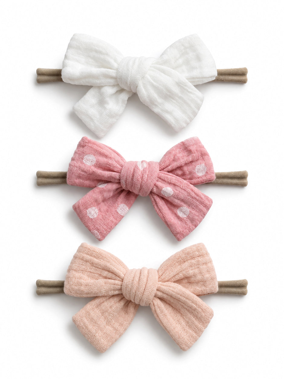 Three hair bows: white, pink with polka dots, and soft pink, all attached to brown hair elastics.