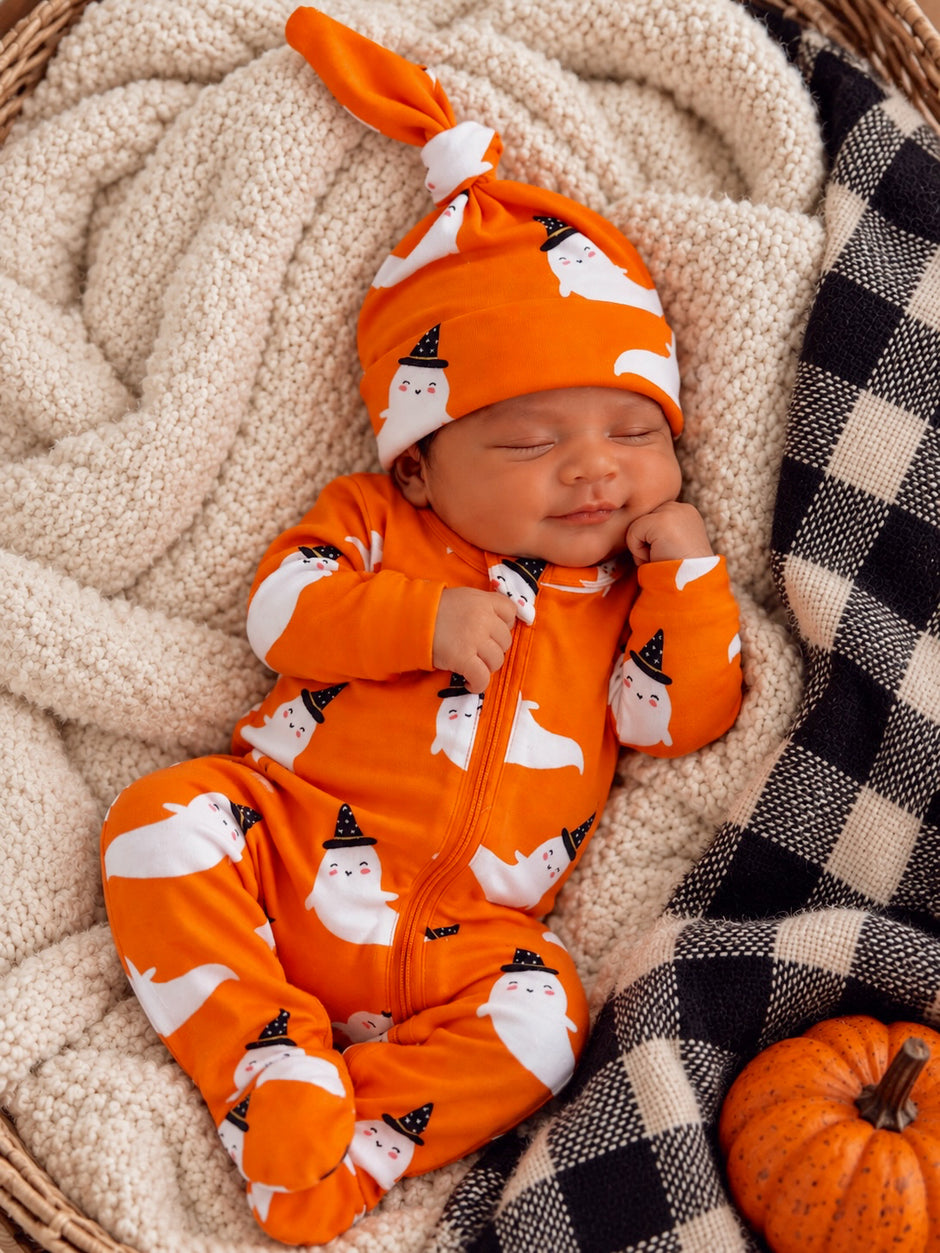 Smiling baby in an orange ghost-themed onesie and hat, resting peacefully on a cozy blanket with a pumpkin nearby.