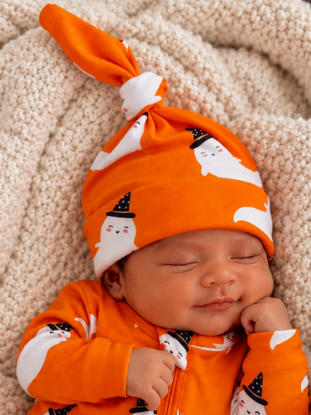 Smiling baby in an orange ghost-patterned hat and outfit, sitting on a soft cream blanket.