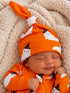 Smiling baby in an orange ghost-patterned hat and outfit, sitting on a soft cream blanket.