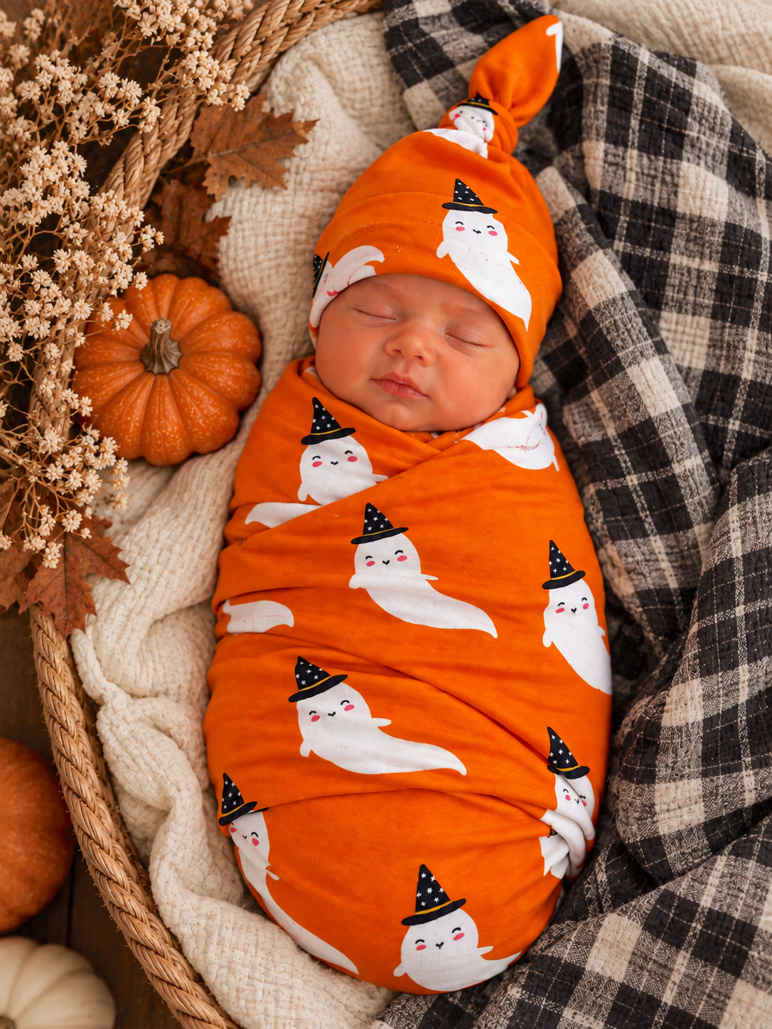 Newborn swaddled in orange fabric with ghost patterns, wearing a matching hat, surrounded by autumn decorations.