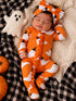 Baby in orange ghost-themed outfit, sleeping on a blanket with pumpkins and a plush ghost toy.