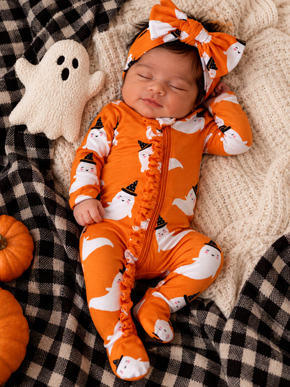 Baby in orange ghost print sleeper with matching headband, resting on a blanket with pumpkins and a ghost plush toy.