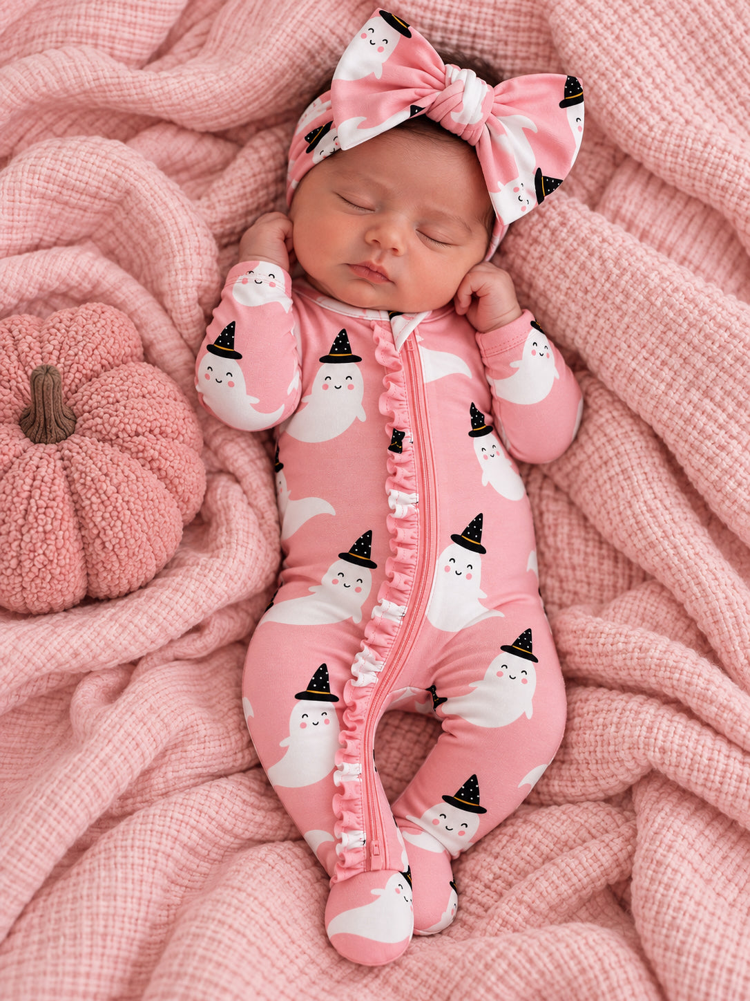 Baby girl in pink ghost-patterned onesie and matching headband, resting on a pink blanket with a pumpkin.