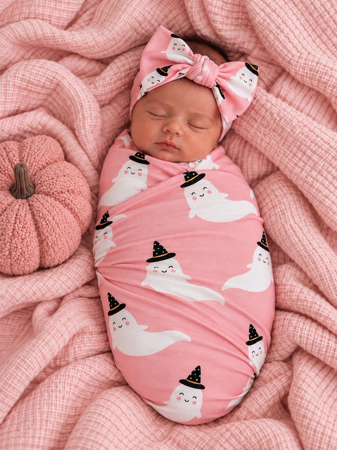 Infant swaddled in pink fabric with ghost prints, wearing a matching headband, surrounded by soft textiles and a pumpkin.