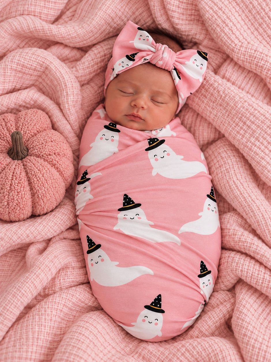 Infant swaddled in pink fabric with ghost prints, wearing a matching headband, surrounded by soft textiles and a pumpkin.