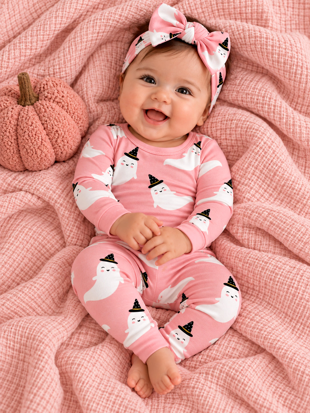 Smiling baby in pink ghost-themed pajamas sits on a soft blanket with a pumpkin, wearing a matching headband.