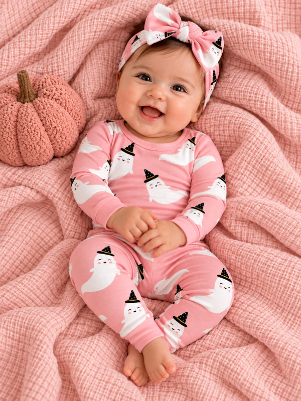 Smiling baby in pink ghost-themed pajamas sits on a soft blanket with a pumpkin, wearing a matching headband.