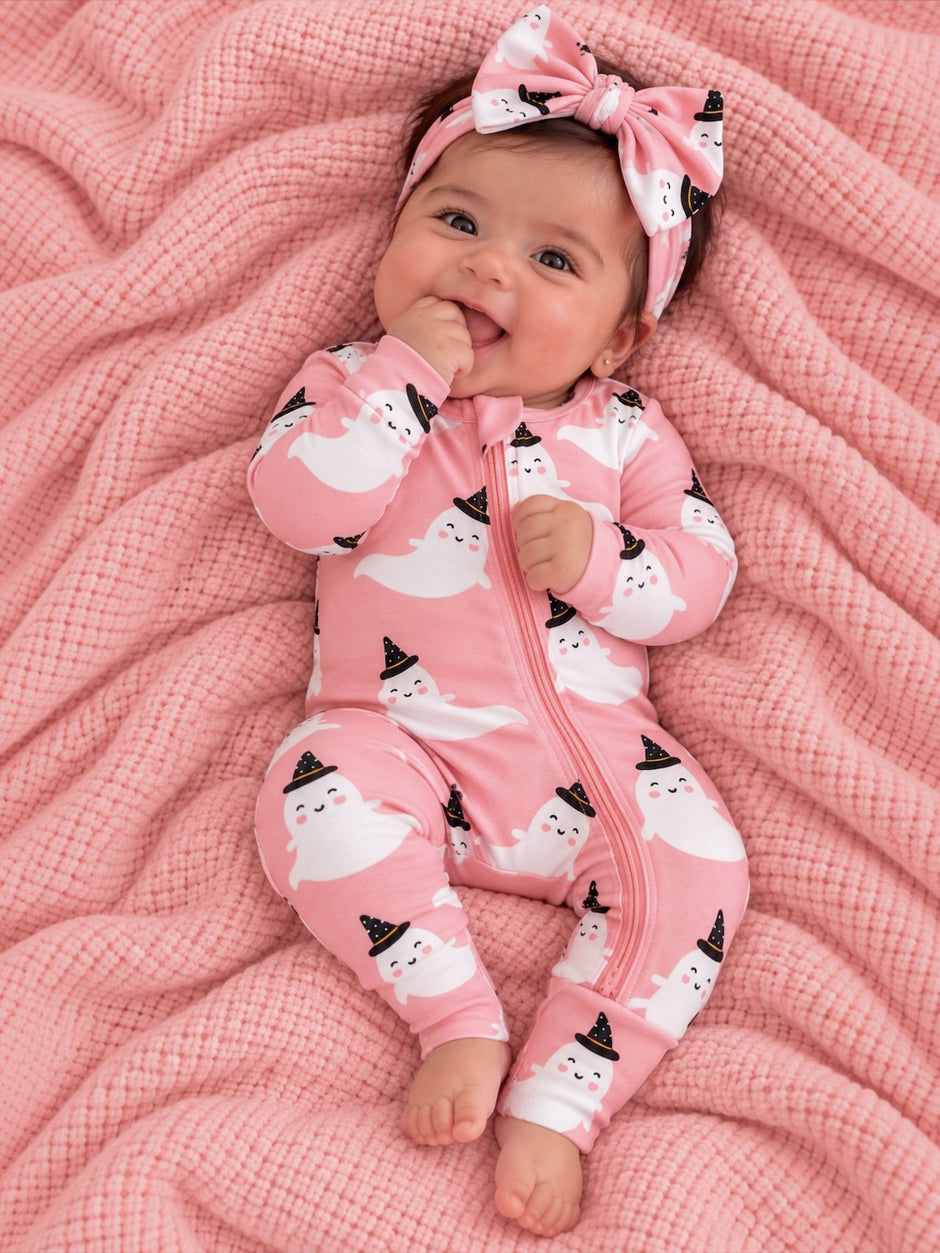 Smiling baby in a pink ghost-printed onesie and bow, lying on a pink blanket.