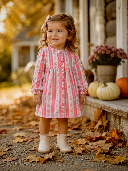 Smiling young girl in a pink floral dress, standing on autumn leaves with pumpkins in the background.