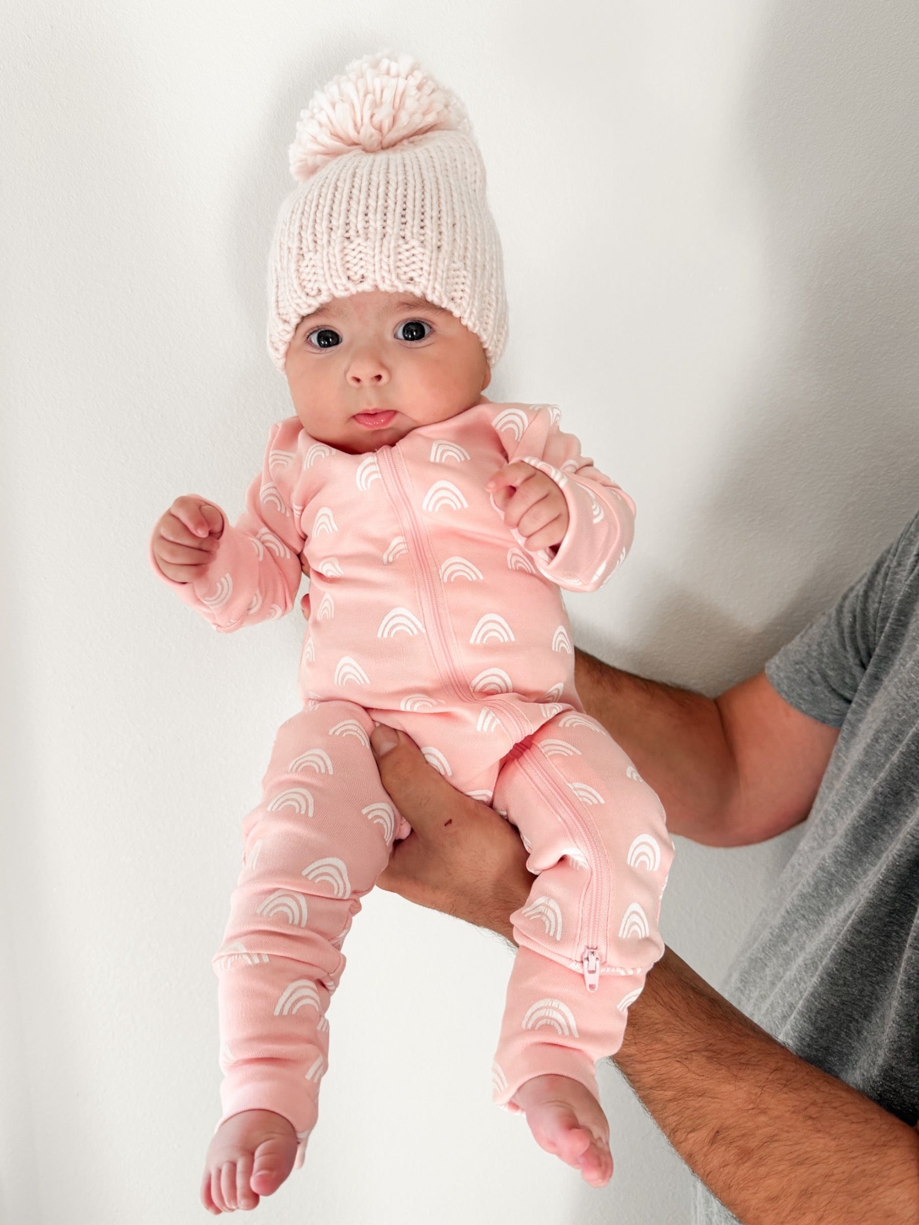 Baby in pink romper with rainbow patterns, wearing a knitted white hat, held by an adult. White background.