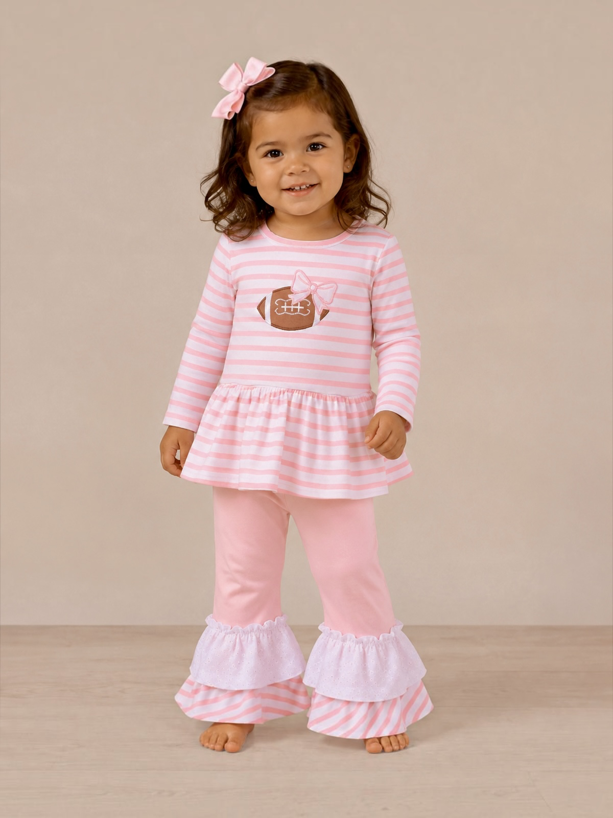 Smiling girl in pink striped outfit with a football graphic and ruffled pants, standing against a neutral background.