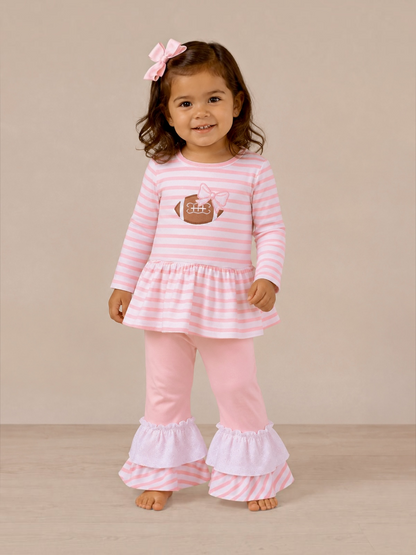 Smiling girl in pink striped outfit with a football graphic and ruffled pants, standing against a neutral background.