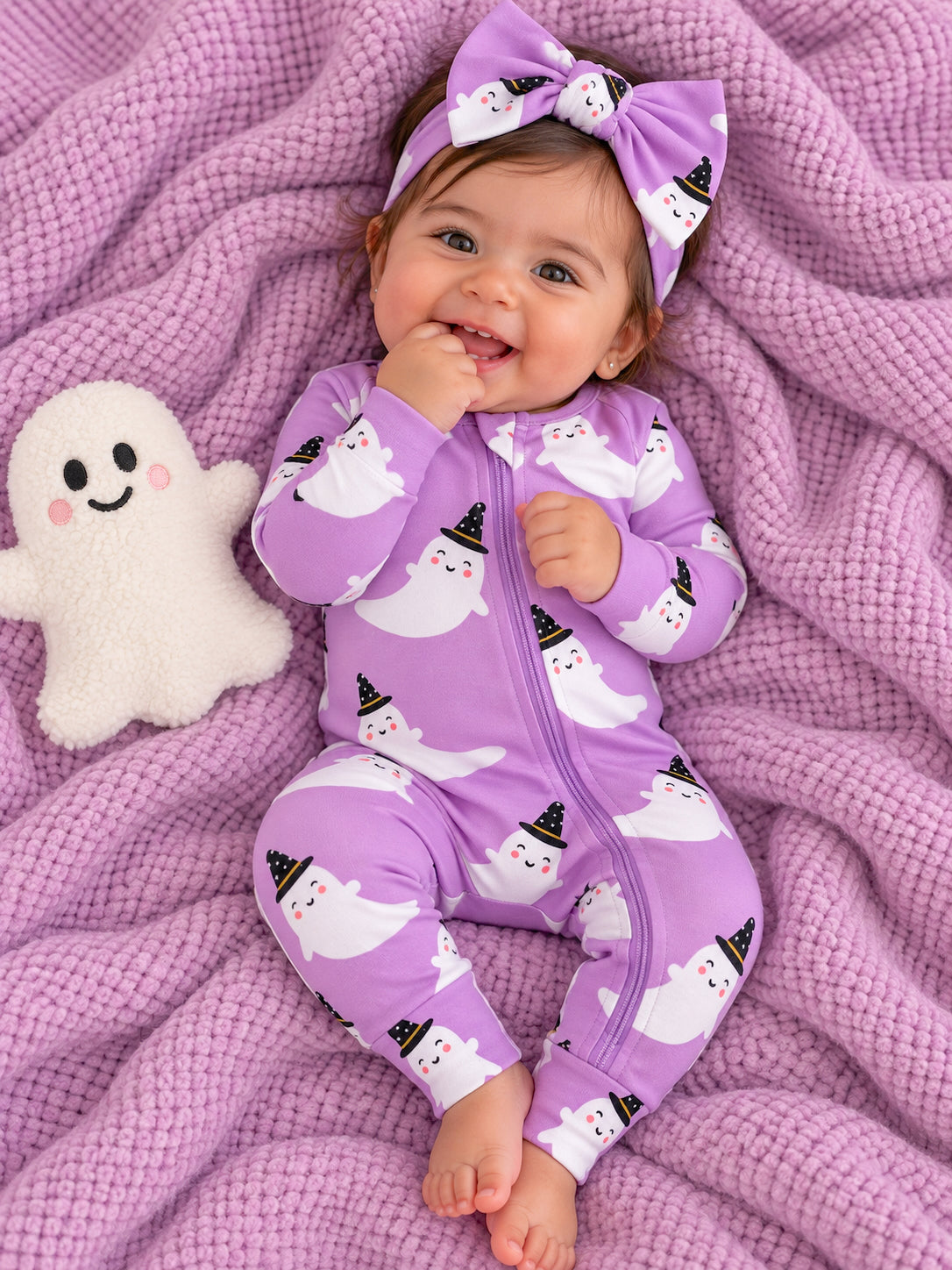 Smiling baby in ghost-patterned purple onesie, surrounded by a soft blanket and plush ghost toy.