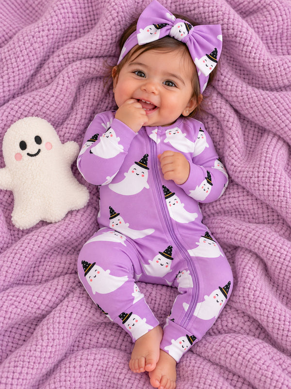 Smiling baby in ghost-patterned purple onesie, surrounded by a soft blanket and plush ghost toy.