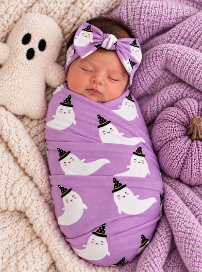 Newborn swaddled in a purple blanket with ghost patterns, next to a plush ghost toy, on a cozy knit backdrop.