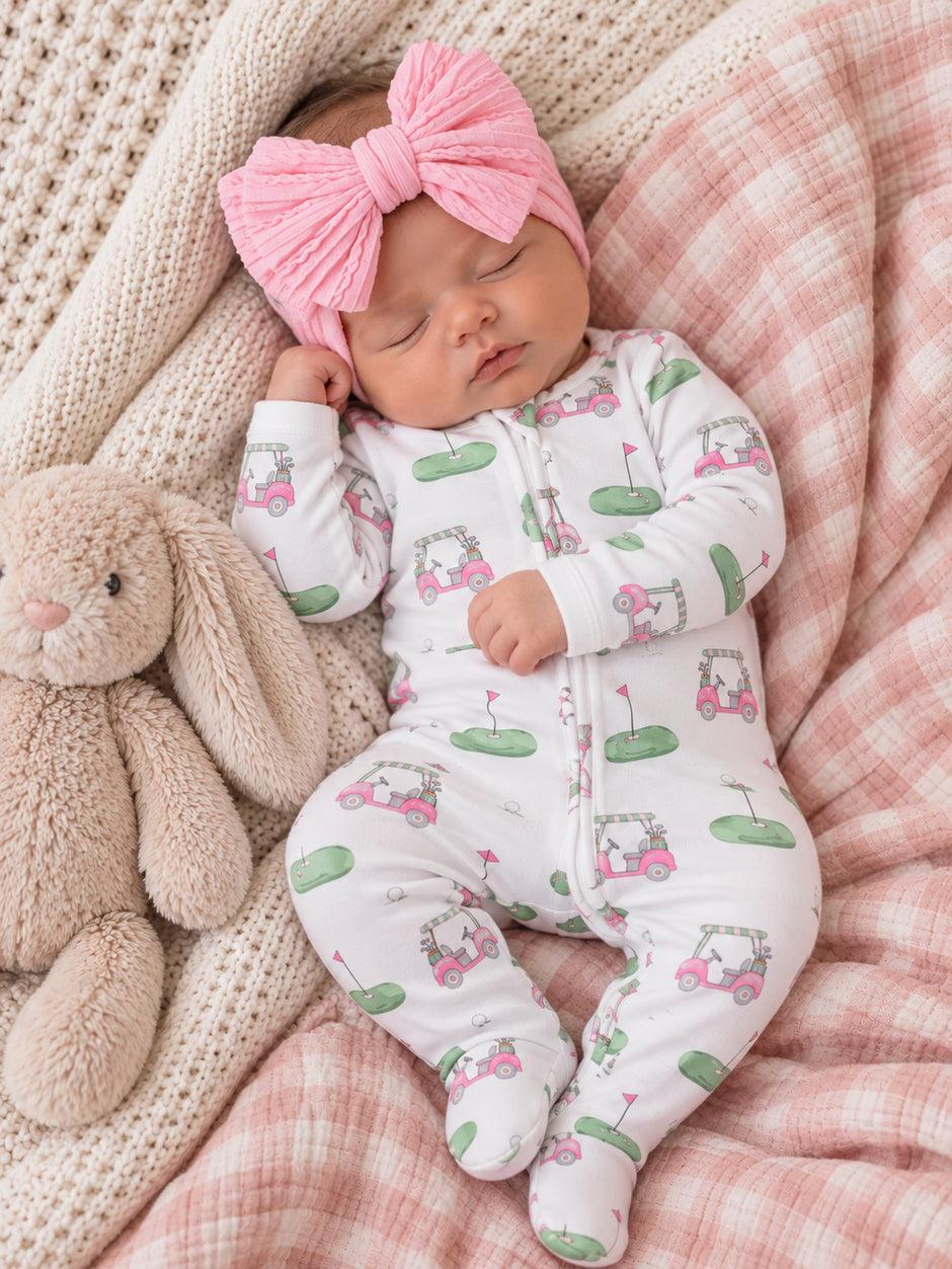 Sleeping baby in golf-themed onesie with pink bow, cuddling a soft bunny on a cozy blanket.