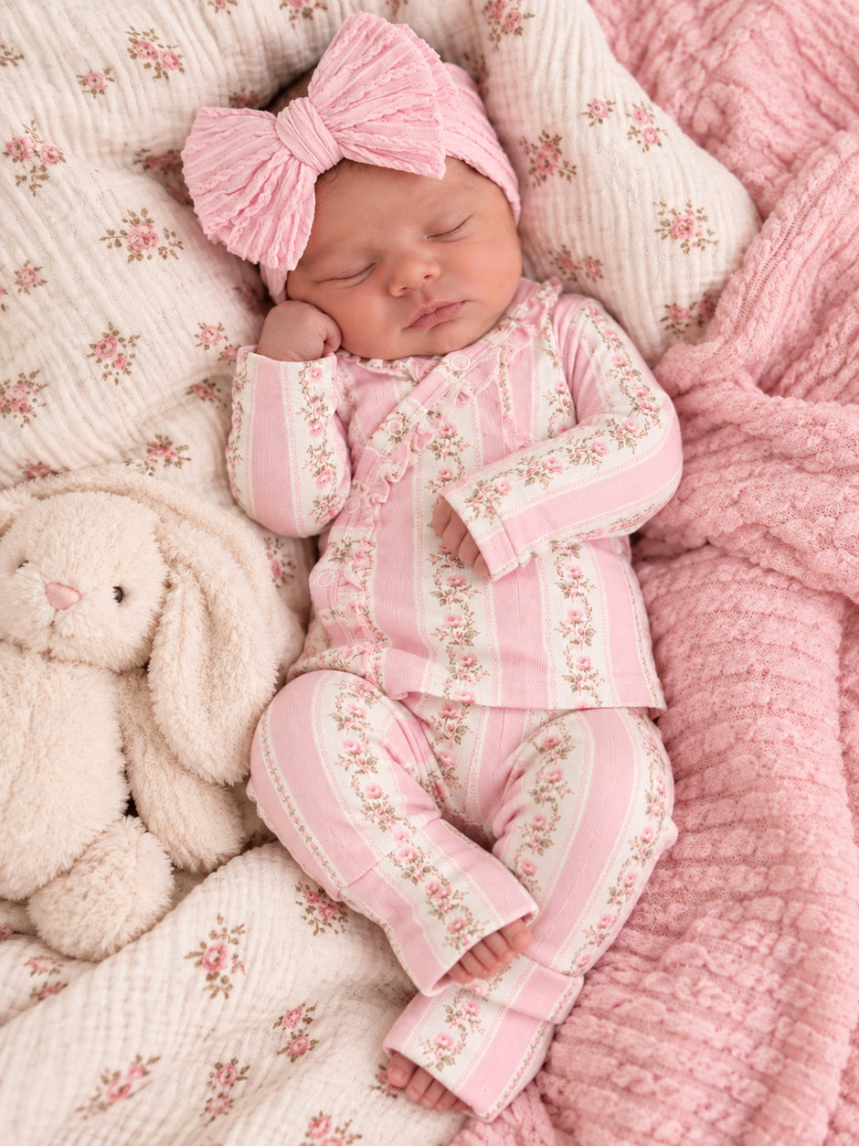 Newborn baby girl in a pink floral outfit with a large bow, sleeping beside a plush bunny on a floral blanket.