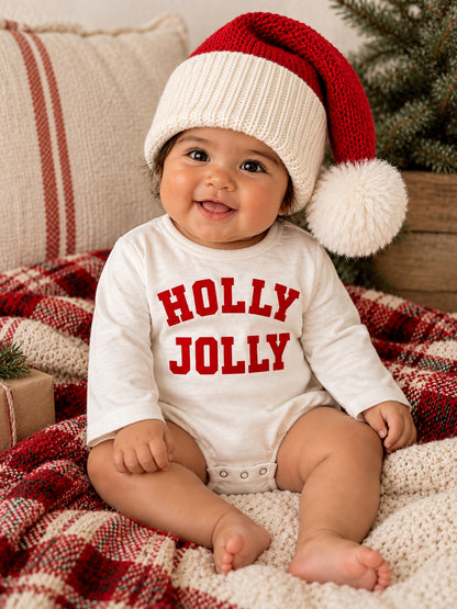 Smiling baby wearing a red and white Santa hat, with a "HOLLY JOLLY" onesie, seated on a cozy plaid blanket.