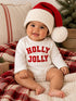 Smiling baby wearing a red and white Santa hat, with a "HOLLY JOLLY" onesie, seated on a cozy plaid blanket.