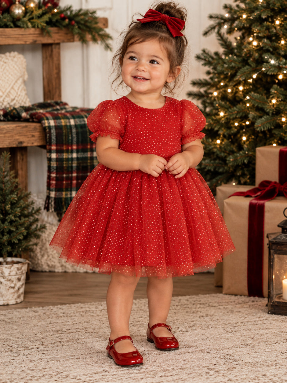 Smiling toddler in a festive red dress with a bow, surrounded by Christmas decorations and gifts.
