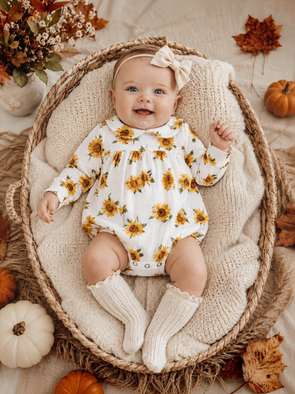 Smiling baby in a sunflower outfit, sitting in a woven basket with autumn decor.
