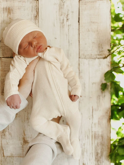 Newborn in a cream-colored outfit and hat, held gently against a rustic wooden background with greenery.