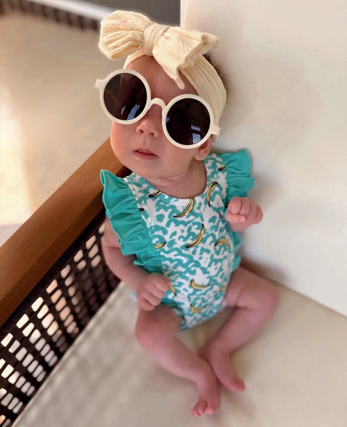 Baby wearing oversized sunglasses and a headband, sitting on a chair in a colorful swimsuit with a banana pattern.