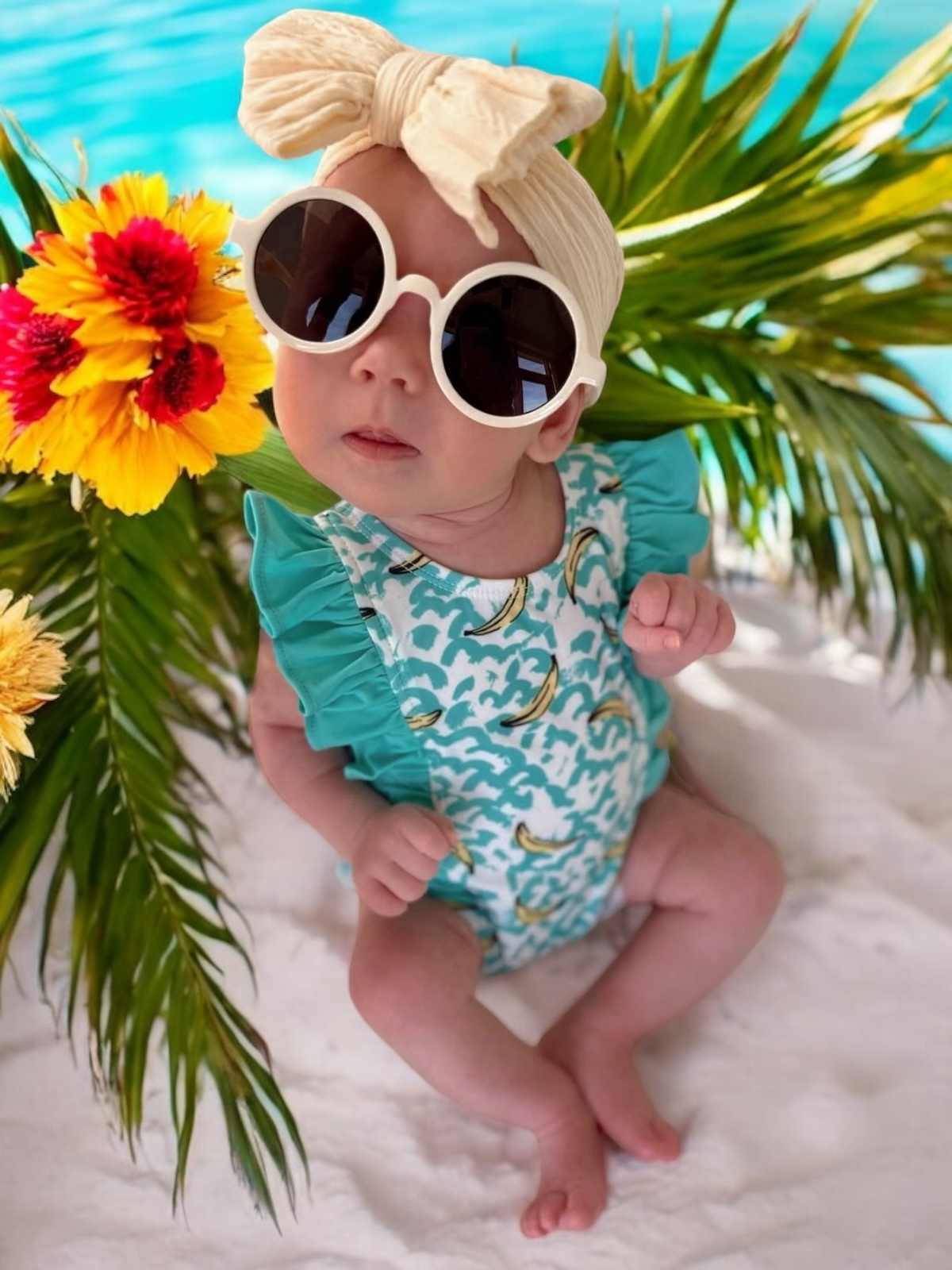 Baby wearing sunglasses and a swimsuit poses with flowers and palm leaves by a pool. Bright and playful summer vibes.