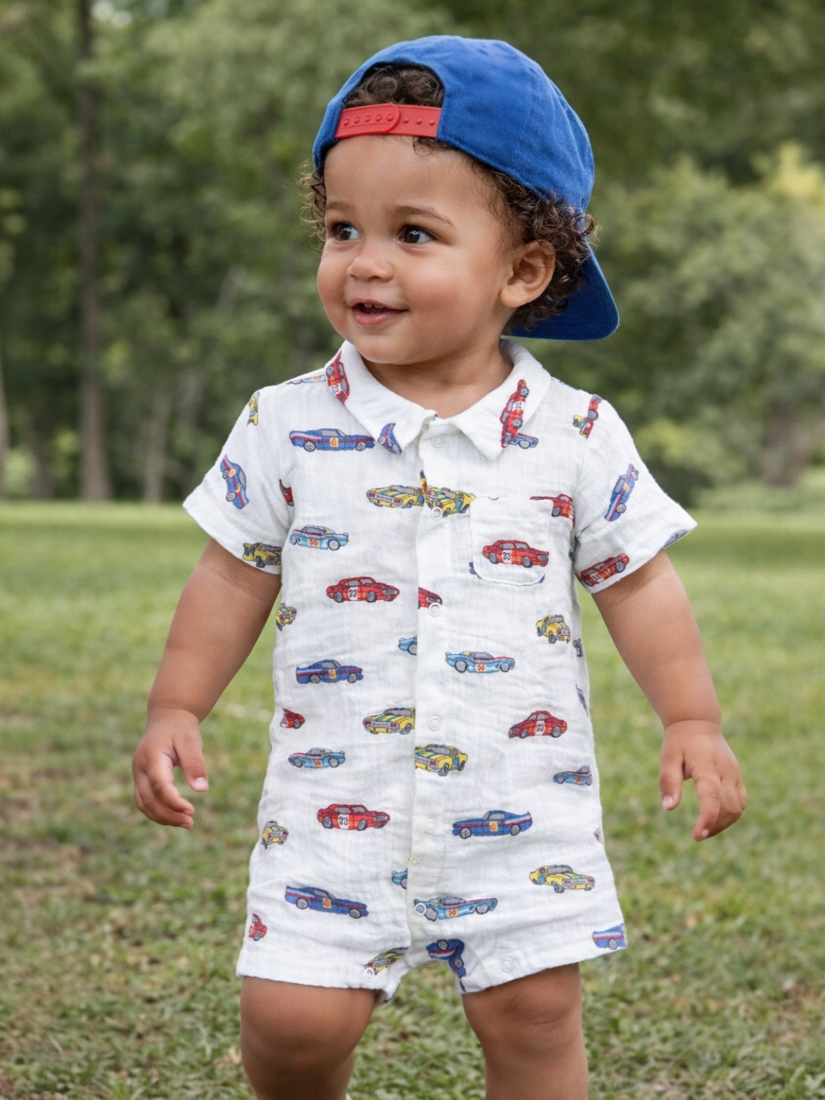 Smiling toddler in a car-patterned outfit and blue cap, standing on grass in a park.