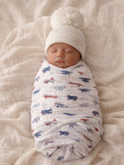 Baby swaddled in a white blanket with airplane patterns, wearing a white pom-pom hat, peacefully sleeping.