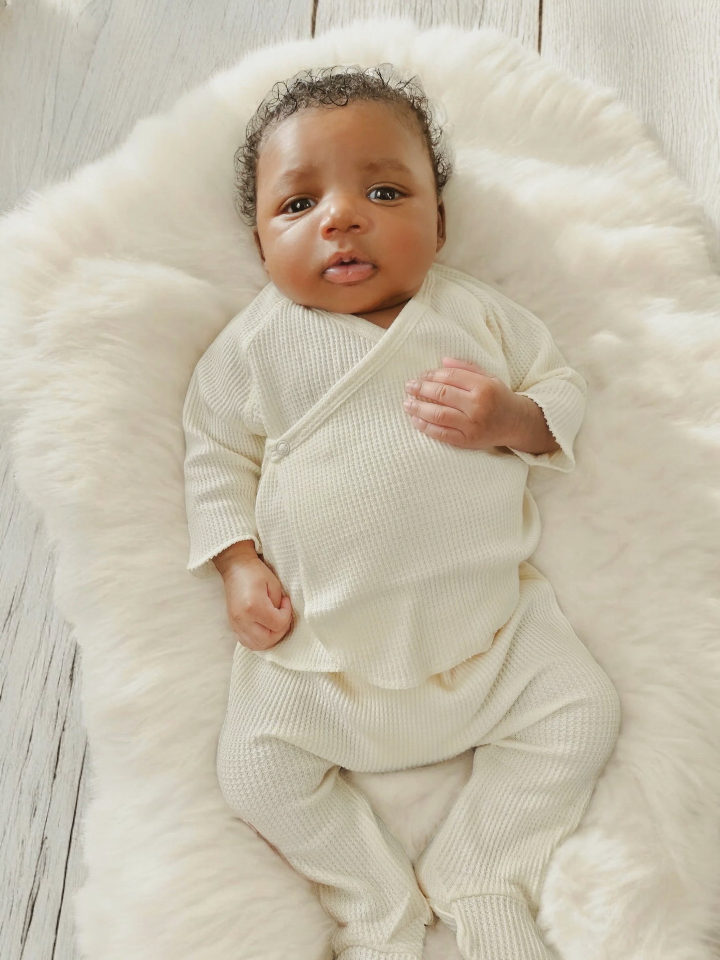Baby lying on a fluffy white blanket, wearing a cream-colored outfit and looking up with a curious expression.
