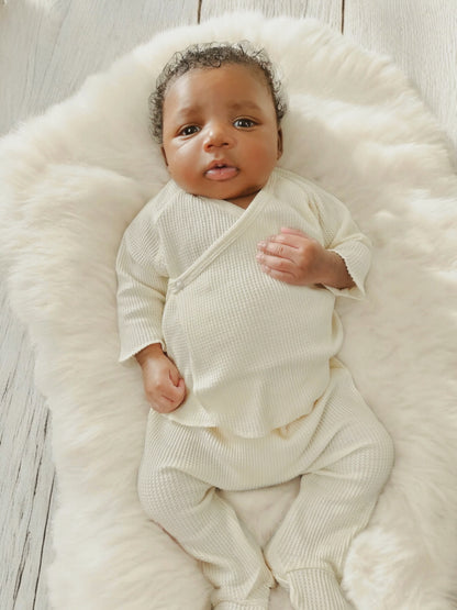 Baby lying on a fluffy white blanket, wearing a cream-colored outfit and looking up with a curious expression.
