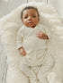 Baby lying on a fluffy white blanket, wearing a cream-colored outfit and looking up with a curious expression.